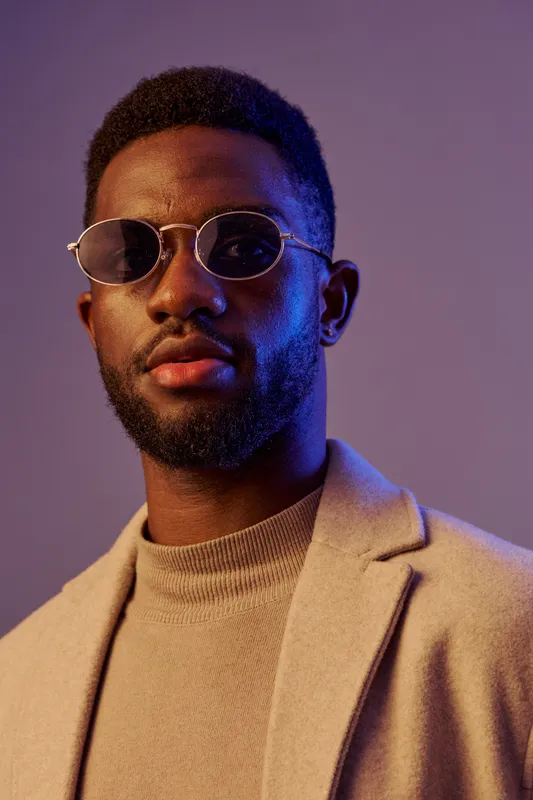 Stylish young Black man wearing trendy sunglasses and a beige coat, exuding confidence in a modern studio setting with dramatic lighting