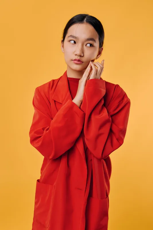 Young woman with long black hair wearing red robe standing against orange background posing thoughtfully with hands near face in casual fashion portrait. Female model looking aside thoughtfully.