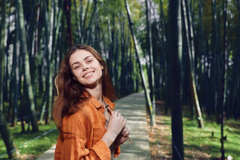 Woman smile forest path backpack nature portrait walking happy traveler among tall trees, orange jacket and relaxed outdoor mood with sunlight, candid portrait of a young hiker in greenery.