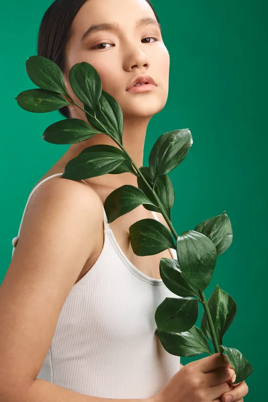 asian woman holds a leafy branch against a vibrant green backdrop, presenting a natural, fresh vibe. she wears a light top, calm and confident, embodying simple beauty and modern style.