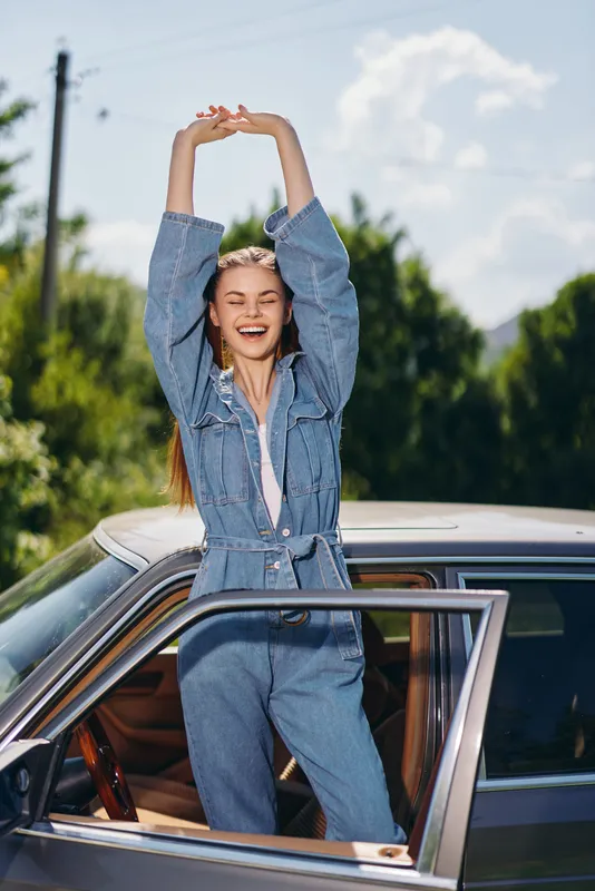 Joyful young woman in denim jumpsuit stretching outside a car in a sunny green landscape, embodying happiness and freedom with an outdoor vibe.