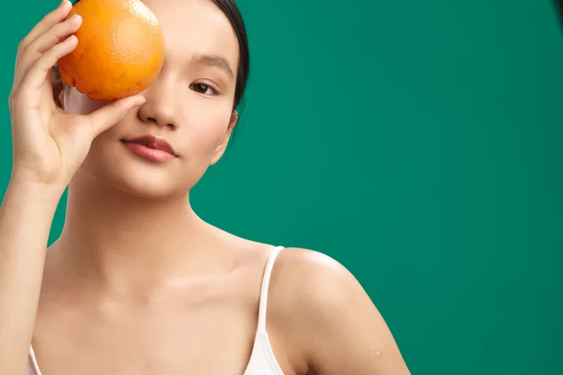 asian woman with a calm expression holds an orange near her eye against a vivid green backdrop, presenting a fresh and playful portrait with bright color contrast and a natural mood.