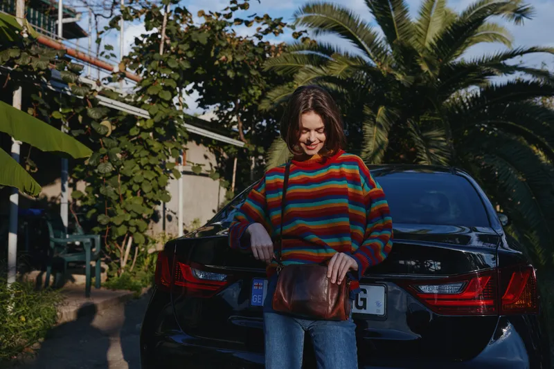 Woman in her 20s, curly brunette wearing a colorful striped sweater and denim jeans, smiling and standing by a black car under palm trees, holding a brown leather bag in sunny tropical driveway.