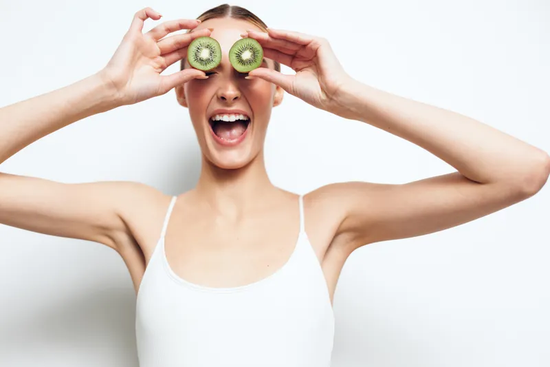 Happy woman holding two kiwi slices over her eyes smiling widely on white background. Concept of healthy lifestyle, fresh fruit, and natural beauty. Bright lighting highlights joyful mood.
