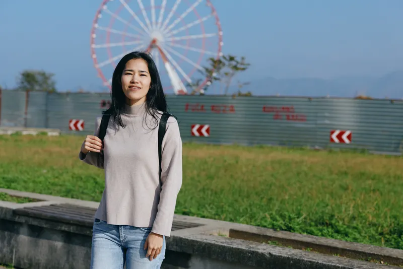 Woman with backpack in a casual sweater and jeans stands in a park with a ferris wheel in the background, conveying travel, leisure and outdoor exploration in daylight