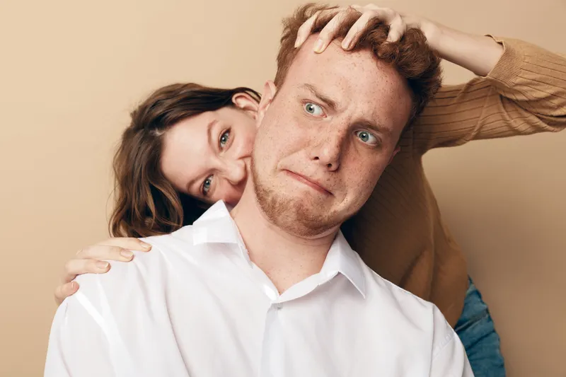 A young woman playfully pulls a young man’s hair while he makes a funny confused face. Both wear casual clothes against a beige background. Concept of humor, relationship, and fun.