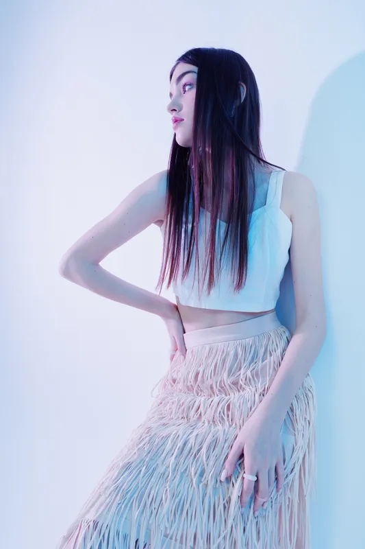 Young woman with long hair wearing a white crop top and fringed skirt poses sideways against a light background. Concept of woman beauty fashion and style.