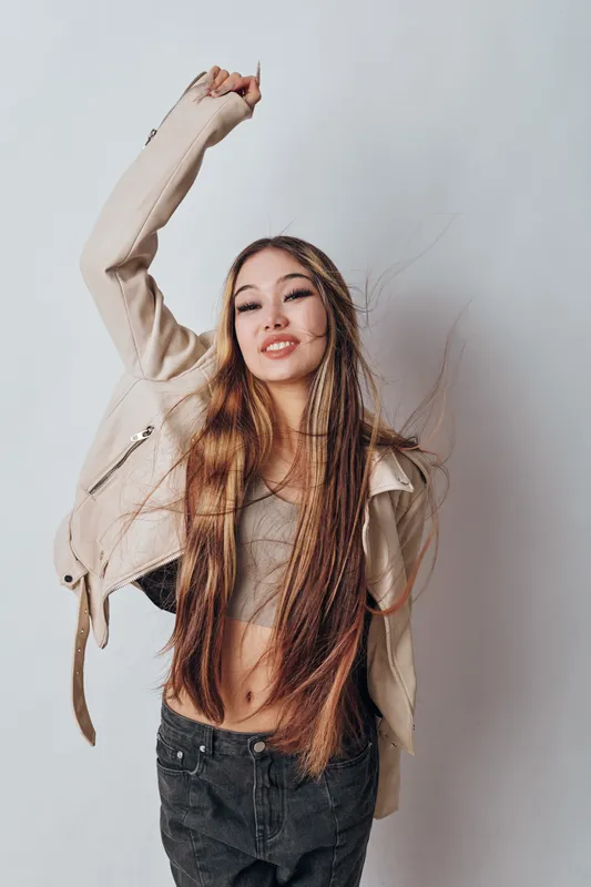 Young woman with long hair wearing casual jacket and crop top posing with raised arm in studio. Smiling female model in trendy outfit with relaxed confident expression and dynamic hairstyle.