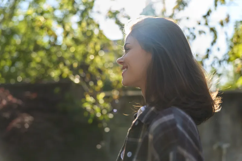 Young woman with shoulder length brunette hair stands in profile outdoors among trees, smiling softly in a casual plaid shirt as warm sunlight filters through leaves on a calm afternoon