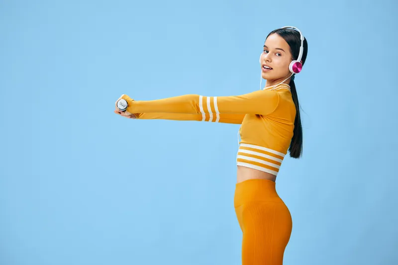 Fitness woman in bright orange athletic wear stretches arms forward, wearing headphones, against a blue studio backdrop, showcasing balance, momentum and athletic readiness.