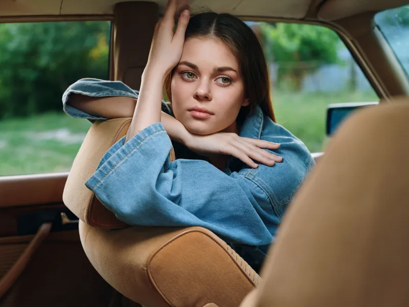 A young woman with long hair in a denim jacket, posing thoughtfully inside a vintage car, showcasing a serene expression in a green natural background.