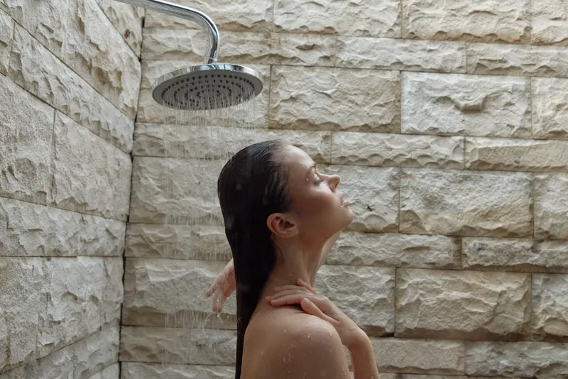 woman taking shower under rain showerhead with exposed stone wall background, wet hair, relaxed expression, natural light highlighting calm moment