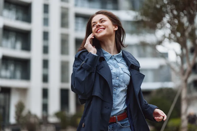 Young woman in blue trench coat talking on smartphone outdoors near modern building, smiling and enjoying conversation. Urban lifestyle, communication, casual fashion concept.