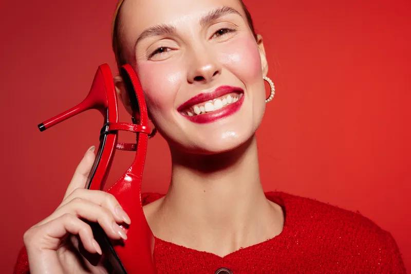 Smiling woman in red sweater holds red high heel shoe near face against red background. Concept of fashion, style, beauty, and confident female model with bright makeup and accessories.
