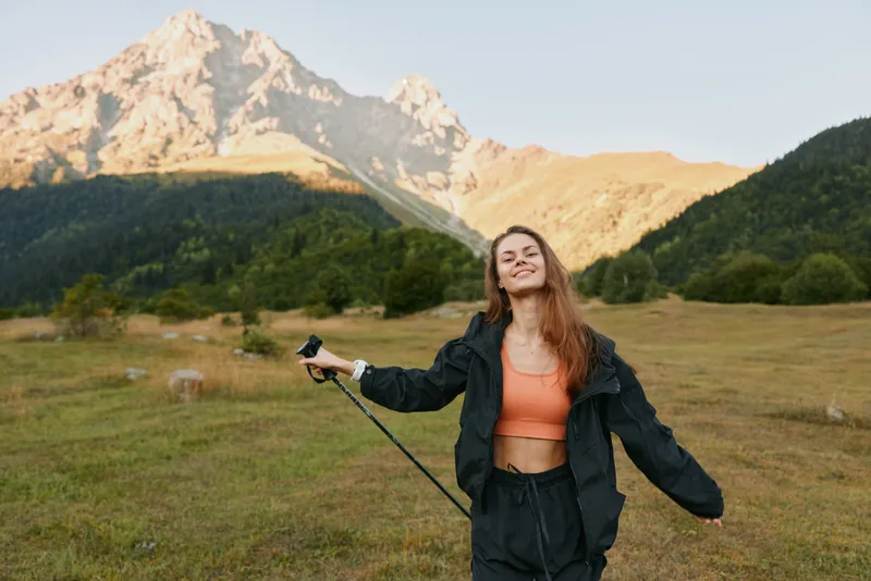 A cheerful outdoor enthusiast stands in a scenic mountain meadow, holding a selfie stick and smiling, enjoying sunny weather and fresh countryside.