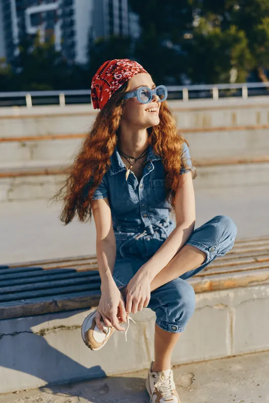 lifestyle woman in modern boho-western denim street style sitting outdoors with sunglasses and red bandana, enjoying sunny day with relaxed, casual vibe in film color