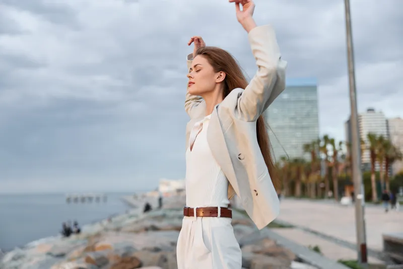 Confident businesswoman in a light suit poses by the seaside, showcasing determination and empowerment against a cloudy sky.