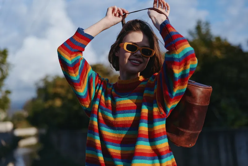 Young woman in rainbow striped sweater and yellow sunglasses smiling outdoors in urban park, carrying brown leather backpack, playful pose adjusting strap, lifestyle fashion travel.