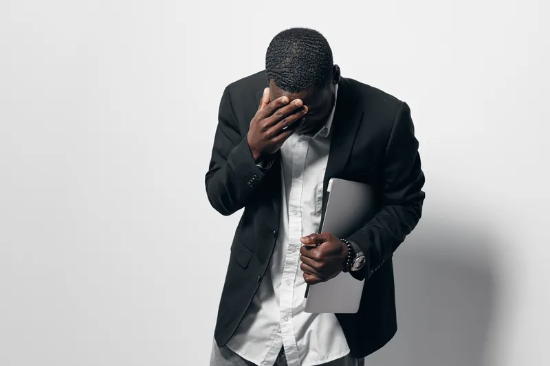 Black man with dark skin in formal black jacket and white shirt holding laptop and covering face with hand, isolated on white background for stress or frustration concept.