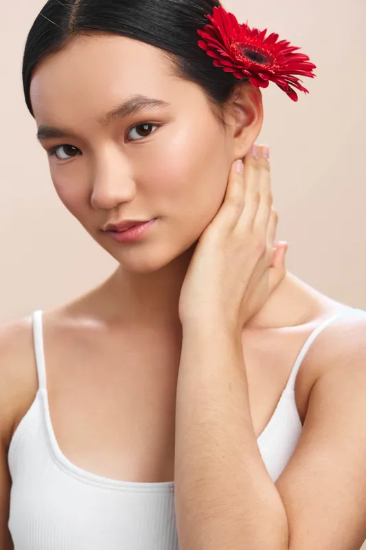 Asian woman with a red flower in her hair gazes softly, showcasing graceful beauty and calm confidence in a simple studio portrait.