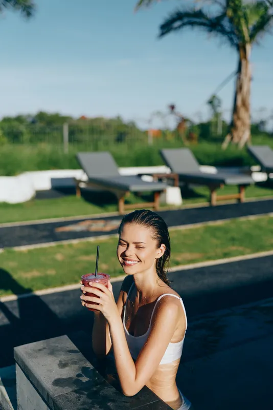 Young woman enjoying a refreshing drink while sitting on the edge of a pool, smiling with closed eyes, sunny day, relaxed atmosphere, tropical outdoor setting.