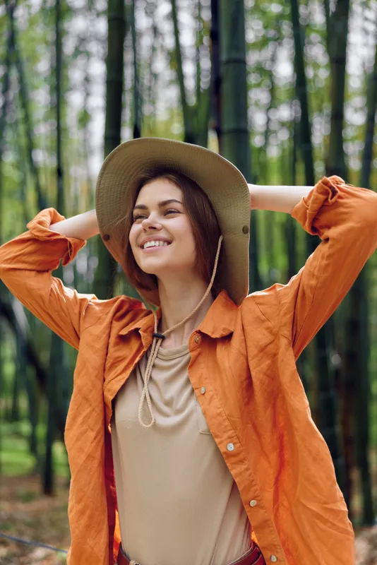 Woman smile hat bamboo forest nature portrait happy travel outdoors: young woman in orange jacket and hat stretches with hands behind head in a bamboo grove, enjoying sunlight and relaxed joyful mood.