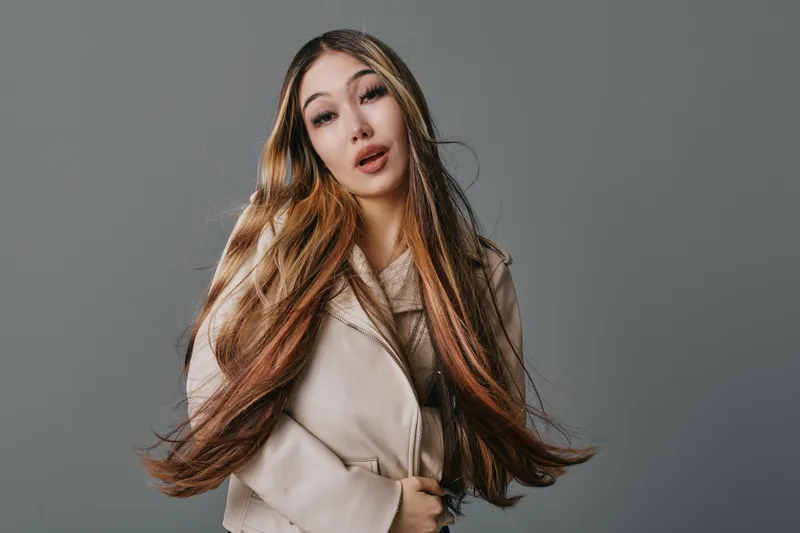 Woman with long brown hair and beige coat posing on grey background. Portrait of young female with neutral expression and hands gently clasping coat for fashion concept.