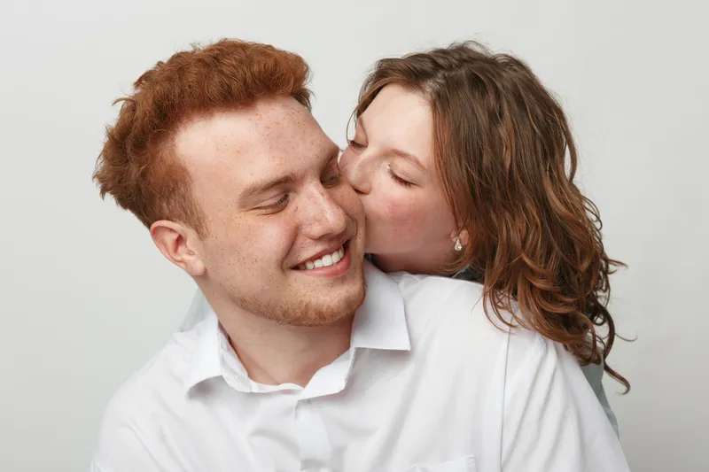Happy young couple in white shirts share affectionate moment, woman kissing man on cheek. Studio portrait on light background. Concept of love, intimacy, and relationship happiness.