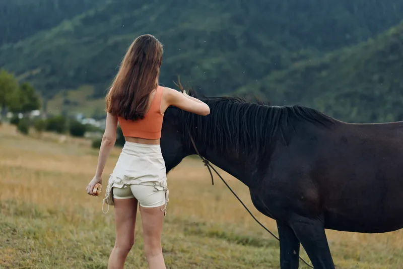 A woman wearing an orange top and light shorts stands beside a dark horse in an open meadow. The scene conveys calm outdoor freedom and gentle companionship in nature.