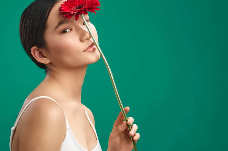 asian woman holds a flower near her face, posing against a teal green backdrop. soft lighting and a calm expression create an elegant, simple portrait with a gentle mood.