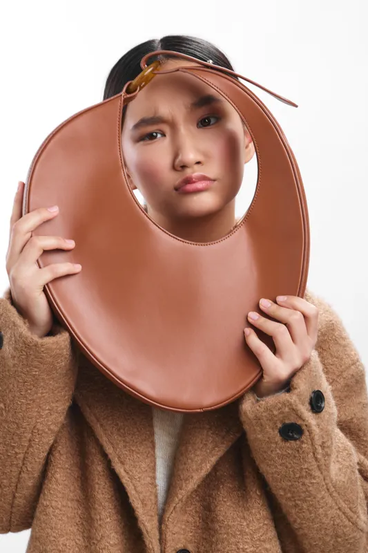 Asian woman wears a cozy coat and holds a large round leather accessory around her head, creating a playful, quirky portrait with bold style.
