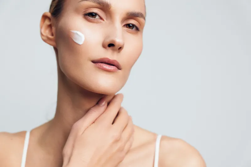 Closeup of young woman applying face cream with gentle hand touch showing smooth skin and natural beauty isolated on light background with soft lighting and calm expression.