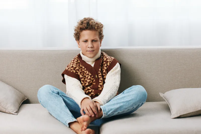A young boy sits cross legged on a sofa wearing a knitted sweater vest and jeans, relaxed expression, natural daylight streaming through a bright living room.