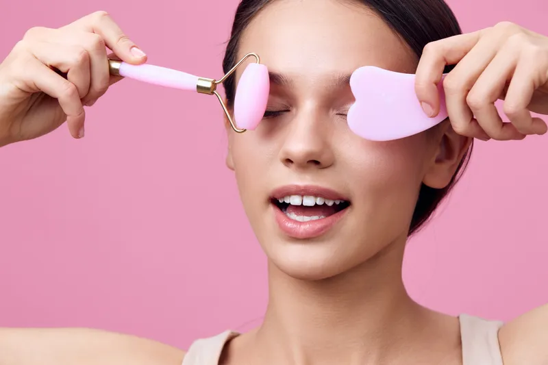 Young woman using facial roller and gua sha tool, enjoying skincare routine on a pink background, promoting beauty and wellness concepts