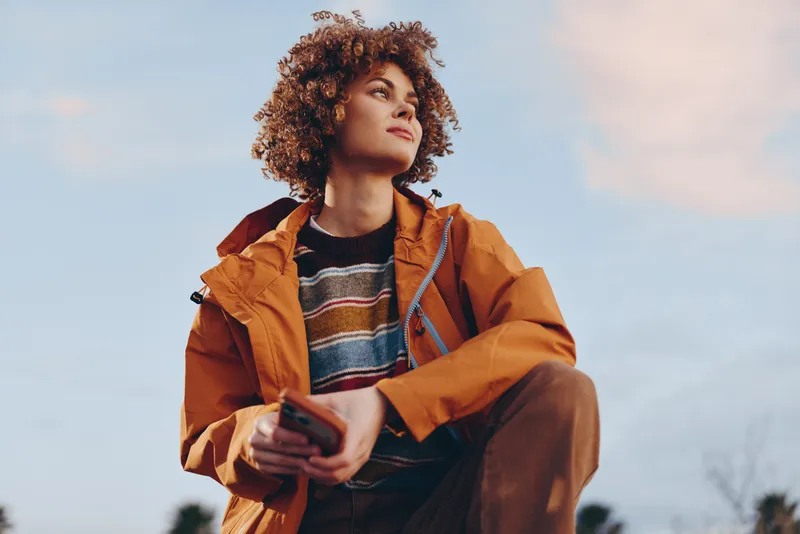Woman outdoor lifestyle curly hair jacket smartphone autumn portrait, young female holding phone while standing in park, warm sunlight, soft wind, relaxed gaze, casual streetwear, sky