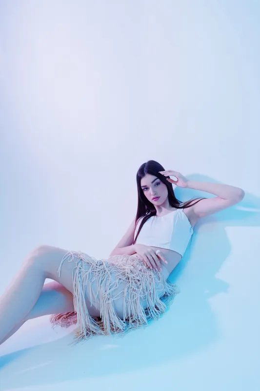 Young woman beauty fashion model in white top and fringed skirt poses sitting on floor with hand near face in soft blue studio lighting. Concept of style and elegance.