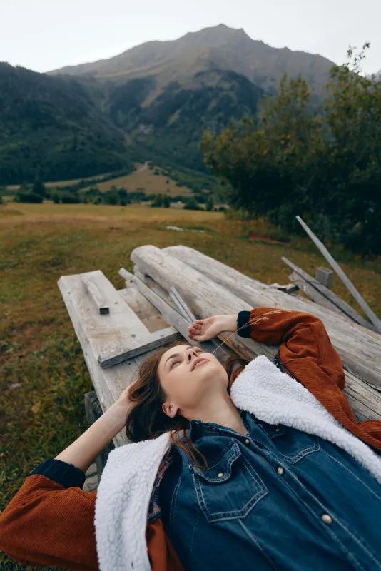 woman relaxation mountains nature meadow outdoors portrait bench: Young woman lies on a wooden bench in a wide meadow, relaxing in a denim jacket and enjoying peaceful mountain landscape and fresh