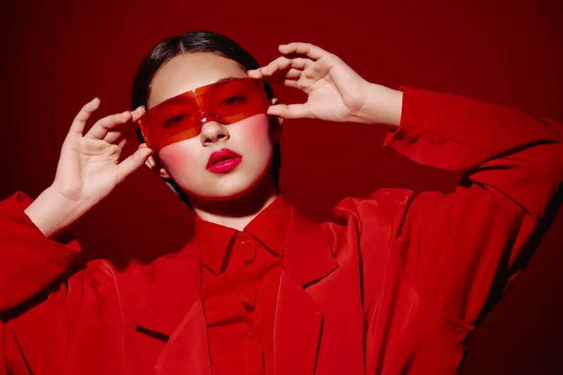 Fashionable young woman wears oversized red coat and matching translucent red sunglasses, posing confidently against deep red background. Concept for beauty, fashion, creative style.