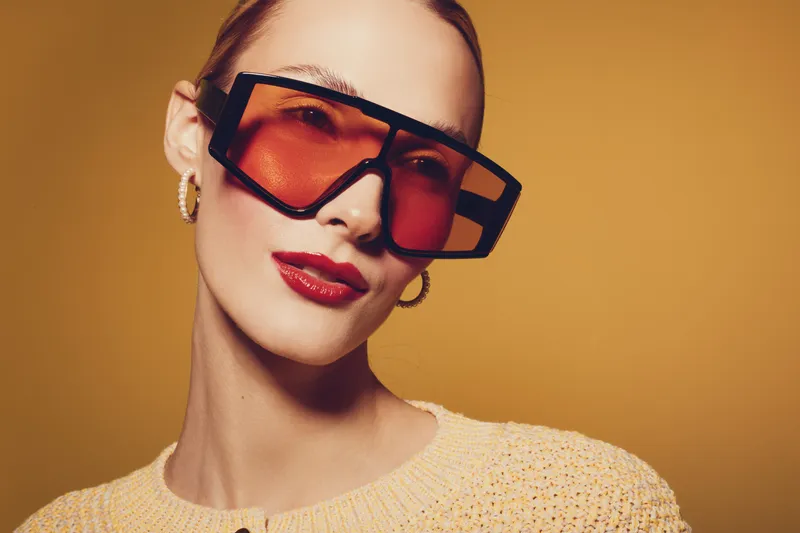 Stylish woman with red lipstick and large orange sunglasses poses against a warm brown background. Fashion concept with modern accessories and confident expression.