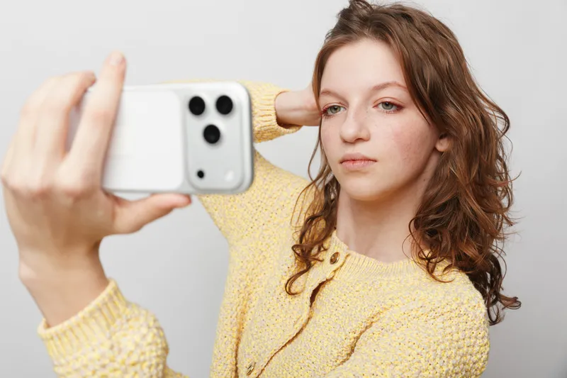 Young woman with curly hair wearing yellow sweater takes selfie with smartphone on white background. Concept of social media, technology, and casual lifestyle portrait.
