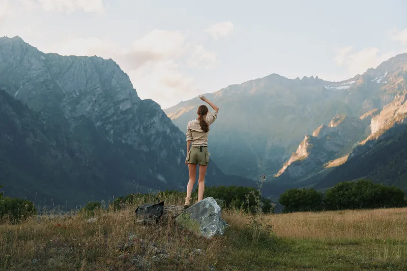 Woman standing on a rock in a grassy field looking at mountain landscape. Outdoor lifestyle scene with natural light and clear sky during daytime.