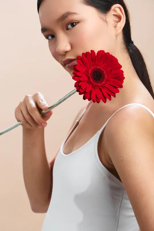 asian woman holds a red gerbera flower near her face in a clean studio, wearing a white tank top. soft lighting highlights her calm expression, creating a graceful, intimate portrait.