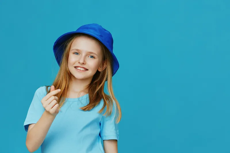 Happy young girl in a blue shirt and hat, showing a heart gesture against a vibrant blue background, symbolizing joy and positivity in childhood moments