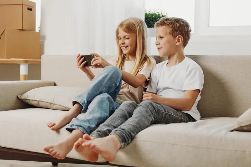 Two smiling children sitting barefoot on a sofa, watching a smartphone together in a bright living room with moving boxes. Concept of family bonding, leisure, and modern childhood.