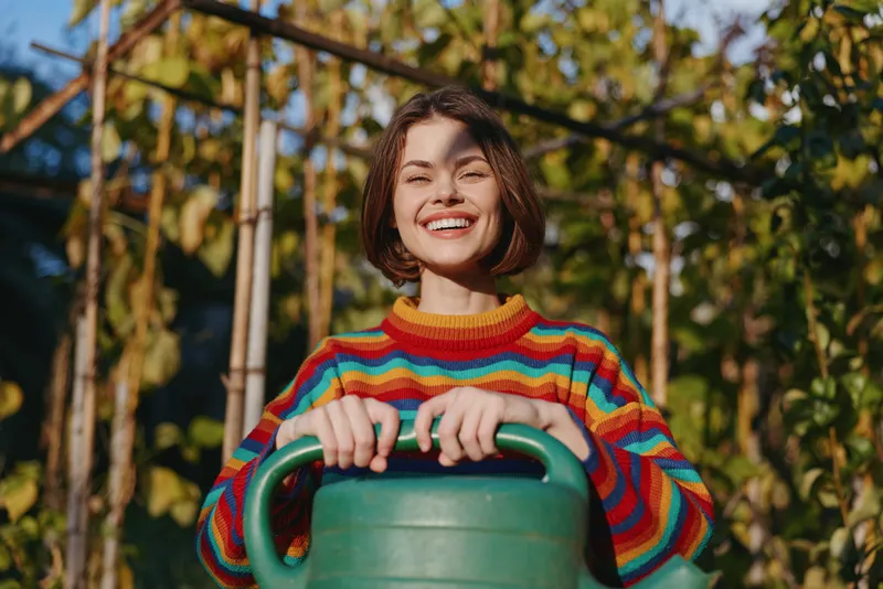 Young adult woman in a colorful striped sweater holding a watering can in a sunlit garden, smiling with short hair, outdoor lifestyle scene for gardening and sustainable hobby.
