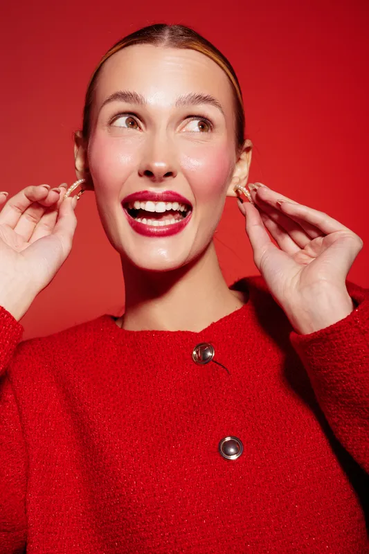 Smiling woman in red sweater adjusts earrings on red background. Close-up portrait with bright makeup and joyful expression. Concept of fashion, beauty, and happiness.