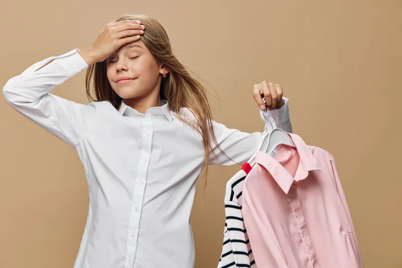 Girl holding two shirts on hangers with closed eyes and hand on forehead standing in front of beige background. Young child deciding between clothes in casual outfit with relaxed expression.