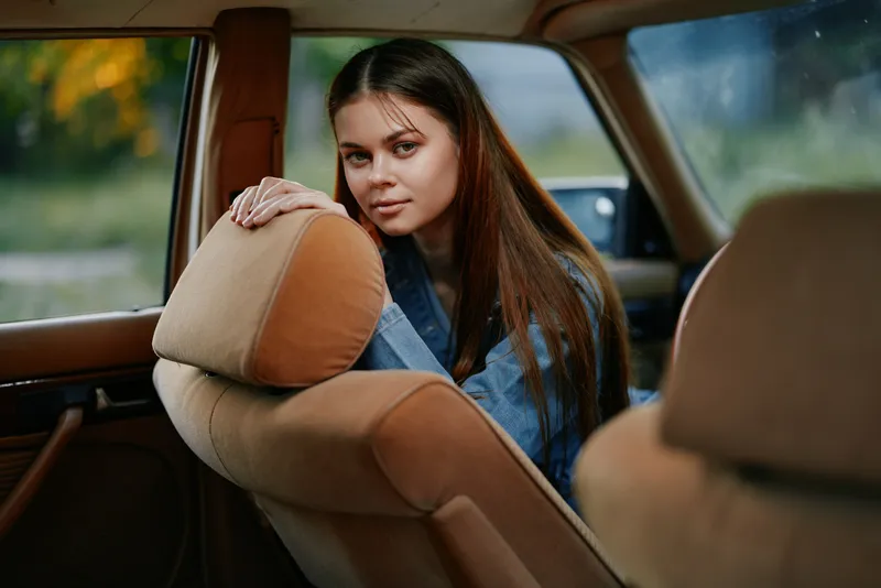 A young woman with long brown hair in a denim jacket sitting in a car's interior, showcasing a relaxed demeanor with a serene background of nature.