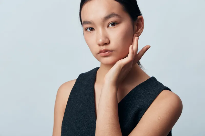 Portrait of young woman with hand on face, natural makeup and contemplative expression on light background. Female model posing in sleeveless top with smooth skin and dark hair pulled back.