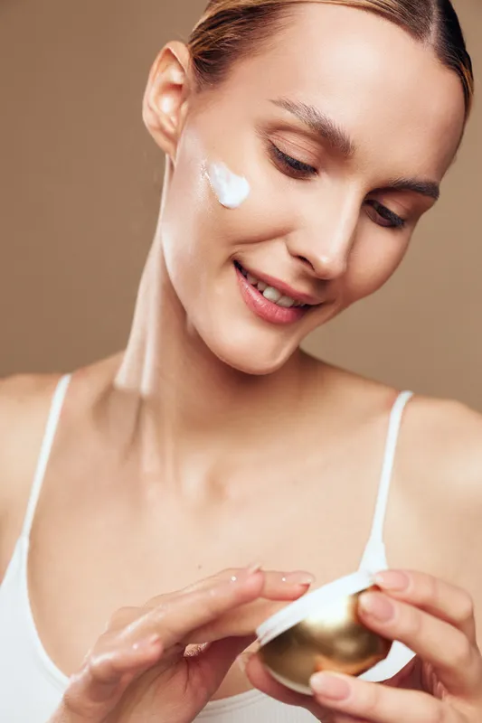 Smiling woman applying white face cream on cheek, holding golden jar. Close-up portrait on beige background. Concept of skincare, beauty routine, wellness, and self-care.